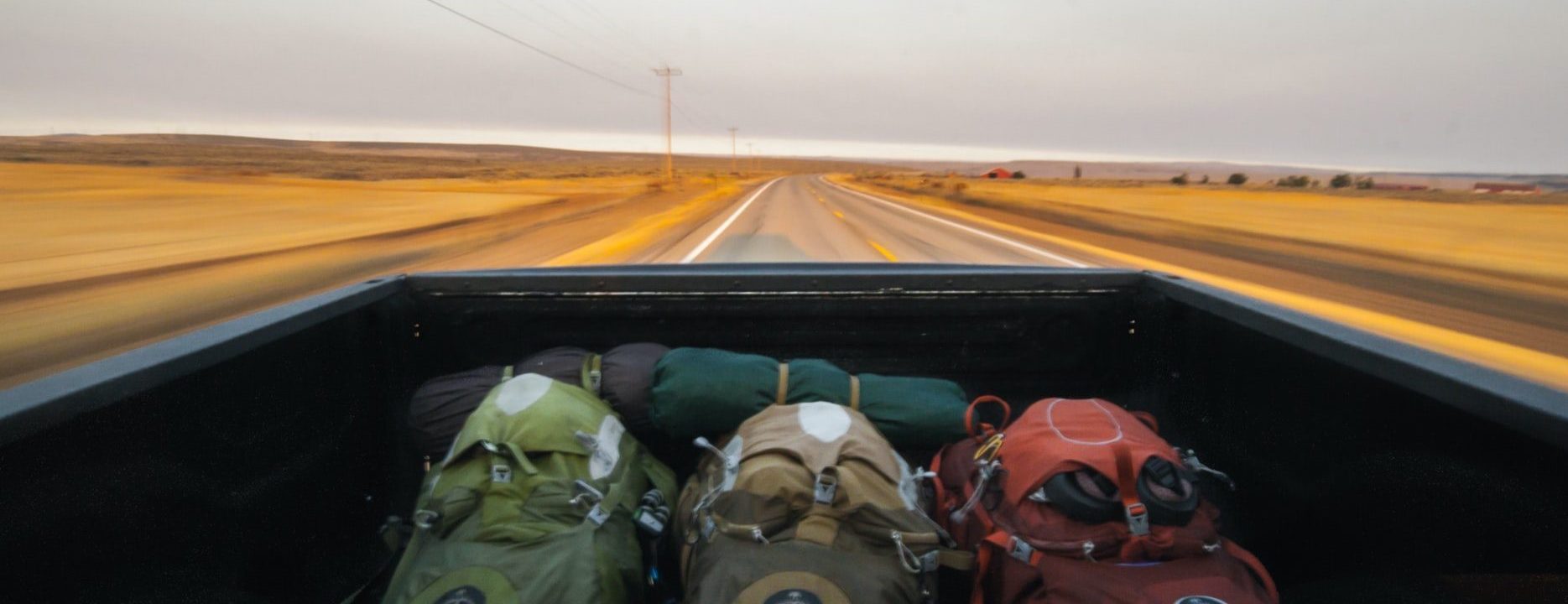 3 zaini in primo piano, appoggiati sul retro di un auto. La foto è scattata dal punto di vista di un passeggero che guarda indietro e vede la strada fatta durante il viaggio.